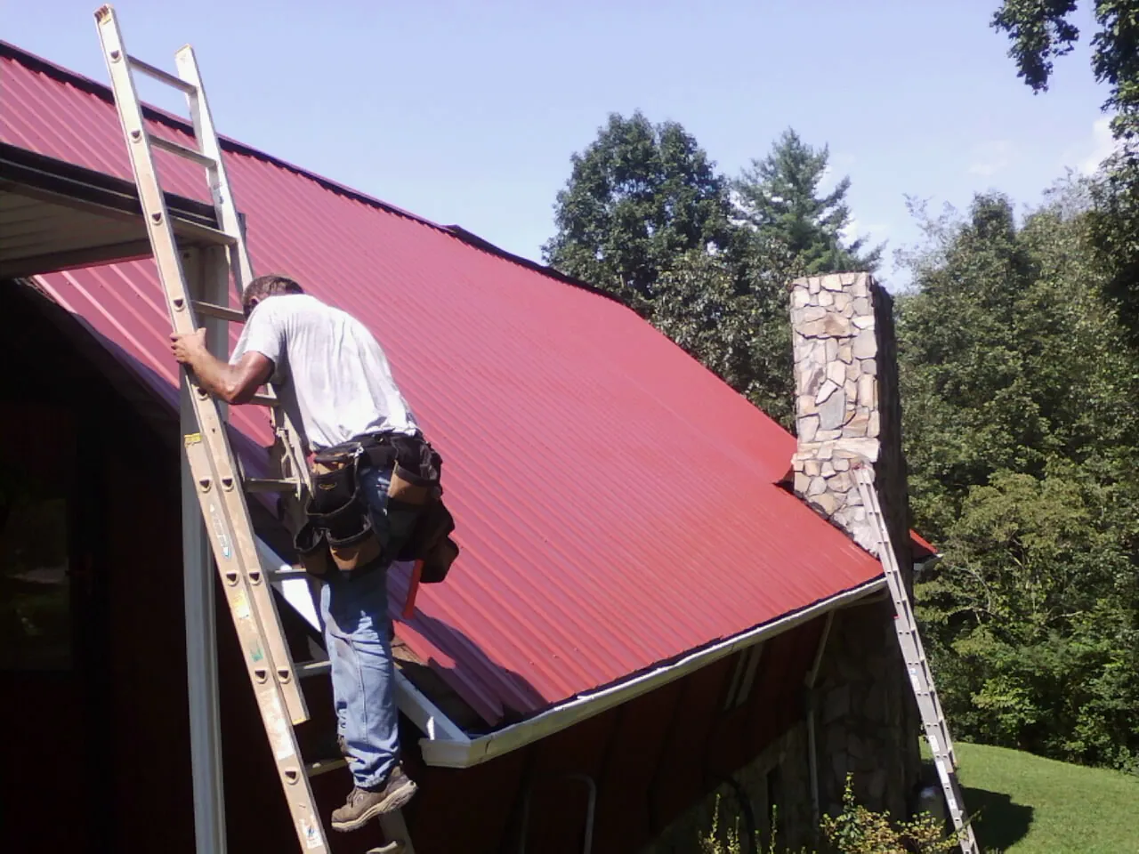 Roofer on a ladder beside a red metal roof and stone chimney — Soffit & Fascia Repair by Roofing Brothers in Lake Hiawatha